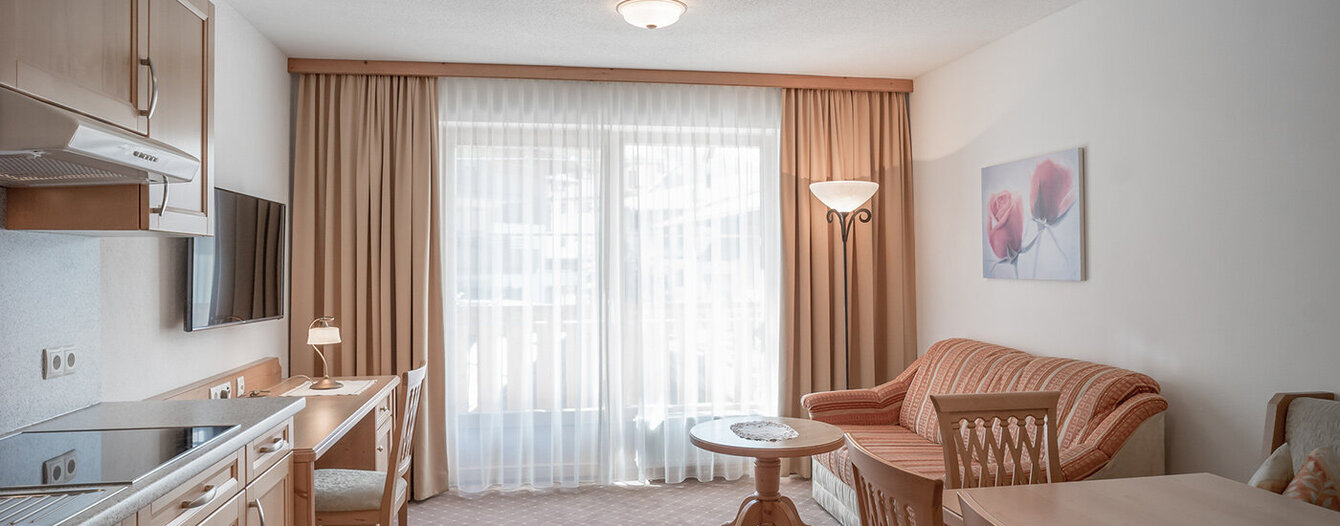 Ein heller, gemütlicher Wohnbereich mit einem Sofa, einem Esstisch und Stühlen aus Holz, einem kleinen runden Tisch, einem an der Wand montierten Fernseher und einer Küchenzeile. Große Fenster mit transparenten Vorhängen lassen natürliches Licht herein. Ein geblümtes Bild hängt an der Wand.