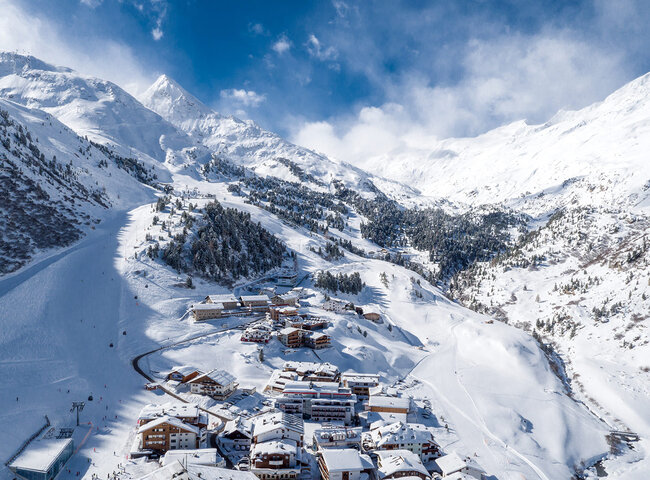 Luftaufnahme von Gurgl mit Skipisten, umgeben von schneebedeckten Gipfeln und Bäumen unter einem teilweise bewölkten Himmel. 