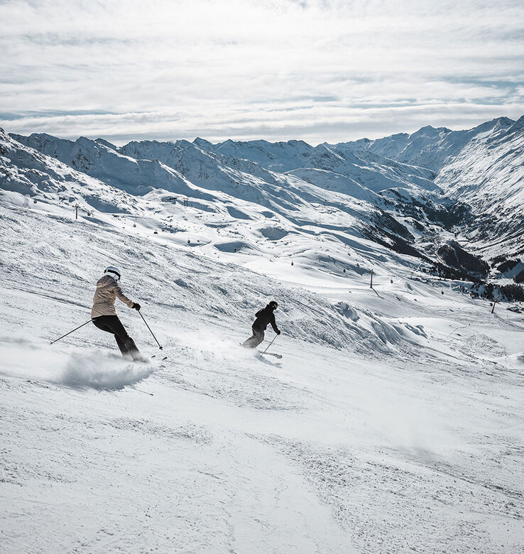 Zwei Skifahrer gleiten einen verschneiten Hang hinunter, der von Bergen umgeben ist. Der Himmel ist teilweise bewölkt, und das Sonnenlicht beleuchtet die Winterlandschaft.