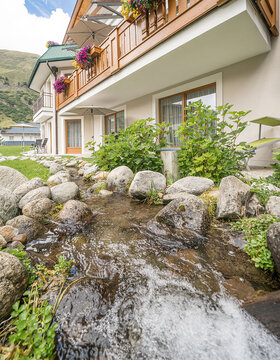 Ein kleiner Bach fließt über Felsen neben einem Gebäude mit Balkonen, die mit bunten Blumen und hölzernen Geländern geschmückt sind, vor grünen Hügeln unter einem teilweise bewölkten Himmel.