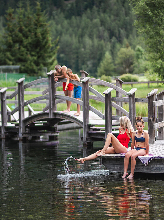 Eine Frau und ein junges Mädchen sitzen auf einem hölzernen Steg und tauchen ihre Füße ins Wasser, während drei Kinder auf einer Brücke im Hintergrund stehen, die von Bäumen und Grünpflanzen umgeben ist.