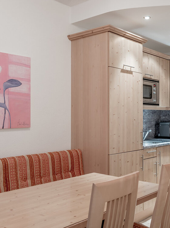 Eine kleine, moderne Küche mit hellen Holzschränken, einer eingebauten Mikrowelle, einem Geschirrspüler und einer Kaffeemaschine. Ein hölzerner Esstisch mit zwei Stühlen und einer gepolsterten Bank steht neben einer Wand mit Blumenkunstwerken.