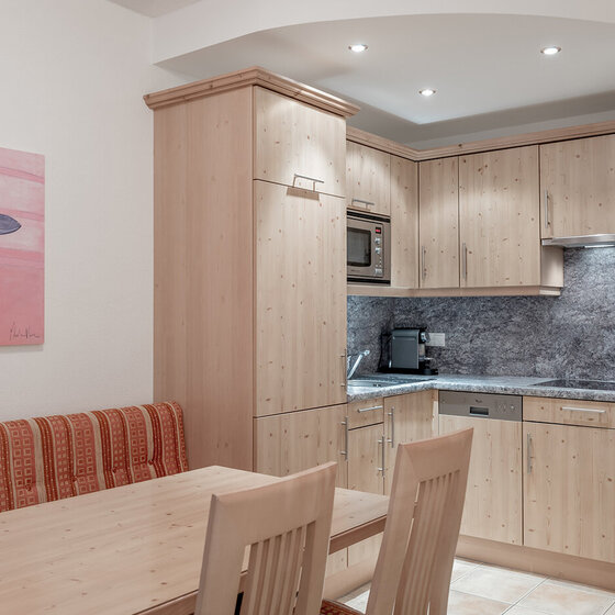 Eine kleine, moderne Küche mit hellen Holzschränken, einer eingebauten Mikrowelle, einem Geschirrspüler und einer Kaffeemaschine. Ein hölzerner Esstisch mit zwei Stühlen und einer gepolsterten Bank steht neben einer Wand mit Blumenkunstwerken.