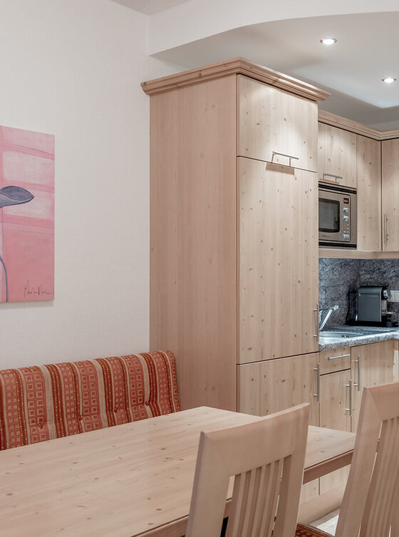 Eine kompakte Küche mit hellen Holzschränken, grauen Arbeitsplatten, Einbaugeräten und einem kleinen Essbereich mit Holztisch, Stühlen und einer orange gestreiften Bank. An der Wand hängt ein geblümtes Gemälde.