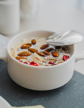 Eine weiße Schüssel mit Müsli, Milch, ganzen Mandeln und Trockenobststücken auf einem grauen Tischset, in der ein Metalllöffel liegt.