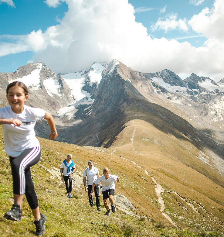 Eine Gruppe von Menschen läuft bergauf auf einem grasbewachsenen Bergpfad mit schneebedeckten Gipfeln im Hintergrund unter blauem Himmel.