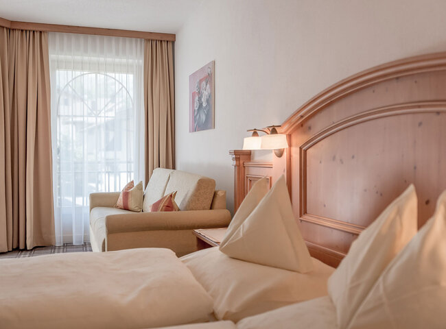 Ein gemütliches Apartmentzimmer mit einem Doppelbett mit cremefarbenem Bettzeug, einem hölzernen Kopfteil, einem beigen Sofa mit roten Kissen, großen Fenstervorhängen, die das Tageslicht hereinlassen, und einem Gemälde an der Wand.