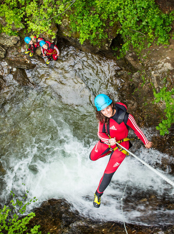 Eine Person in einem roten Neoprenanzug und einem blauen Helm seilt sich lächelnd einen felsigen, rauschenden Wasserfall hinunter.