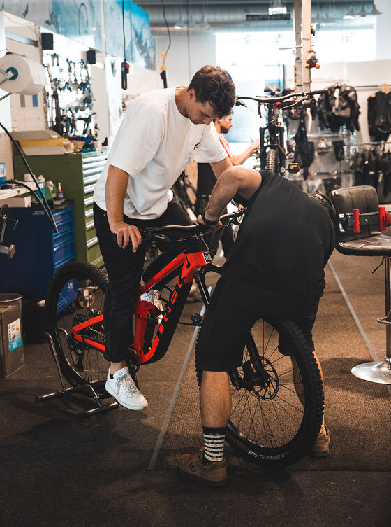 Zwei Personen arbeiten in einer Werkstatt an einem roten Mountainbike. Eine Person sitzt auf dem Fahrrad, während die andere über das Rad gebeugt das Vorderrad repariert. Im Hintergrund sind Werkzeuge und Fahrradteile zu sehen.
