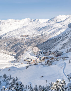 Ein verschneites Alpendorf liegt in einem Tal, umgeben von Bergen, Kiefern und frisch verschneiten Hängen unter einem strahlend blauen Himmel. Winterliches Lagenbild von Gurgl mit dem Alpenresidenz Logo in der Mitte des Bildes.