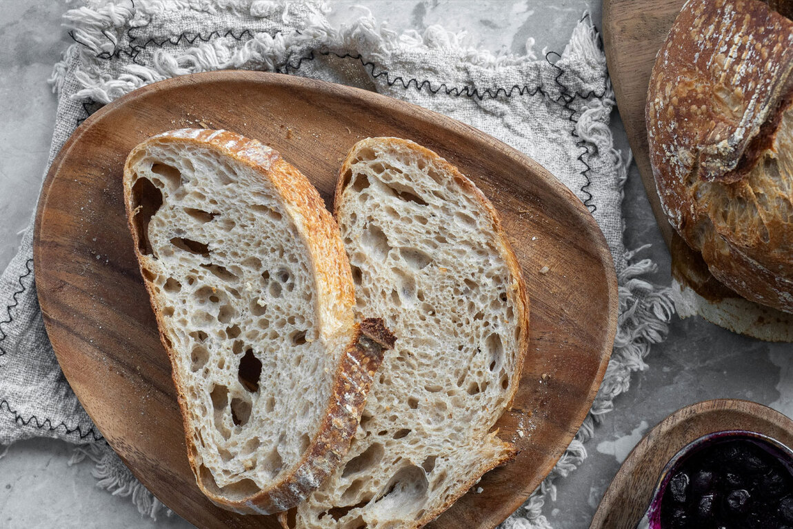 Zwei Scheiben rustikales, luftiges Sauerteigbrot liegen auf einem Holzbrett auf einem strukturierten grauen Tuch, daneben sind ein Teil eines Brotes und eine kleine Schale mit dunkler Marmelade zu sehen.