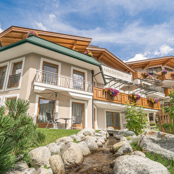 Ein charmantes Alpengästehaus mit Holzbalkonen, bunten Blumenkästen und einem grünen Dach, umgeben von üppigem Grün und Felsen mit einem kleinen Bach im Vordergrund unter einem strahlend blauen Himmel.