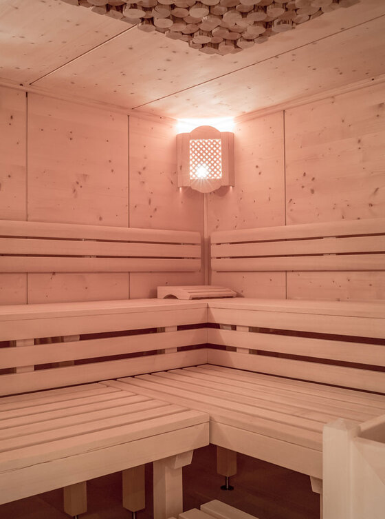 Eine moderne Holzsauna mit L-förmig angeordneten hellen Holzbänken, einem beheizten Steinofen in der Ecke und einer weichen, warmen Beleuchtung an der Wand über dem Ofen.