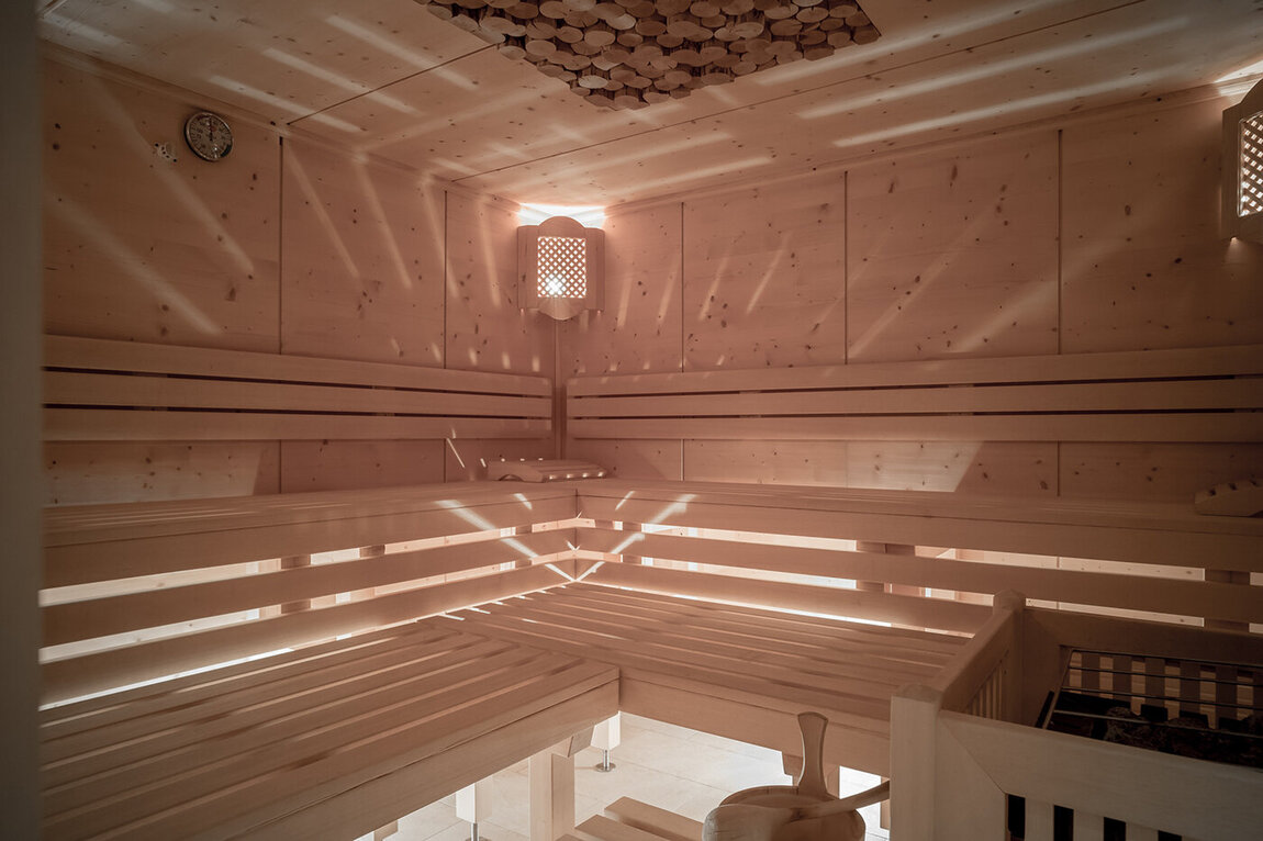 Eine leere Holzsauna mit gestuften Sitzbänken, einer Wanduhr, einem Ofen mit Steinen und sanftem Licht.