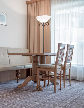 Ein Holztisch mit zwei passenden Stühlen und einer gepolsterten Bank steht in einem hellen Raum mit großen Fenstern, transparenten Vorhängen, einer Stehlampe und einem weichen Teppichboden. Warme, neutrale Töne schaffen eine gemütliche, einladende Atmosphäre.