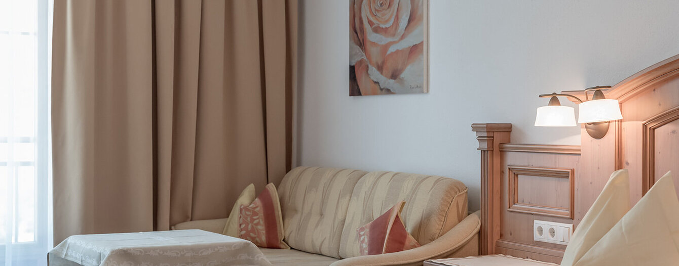 Ein gemütliches, beigefarbenes Apartmentzimmer mit einem Doppelbett, einem kleinen Sofa mit Kissen, einem Holztisch, einem Wandbild mit einer Rose und sanfter Beleuchtung neben einem Fenster mit langen beigen Vorhängen.