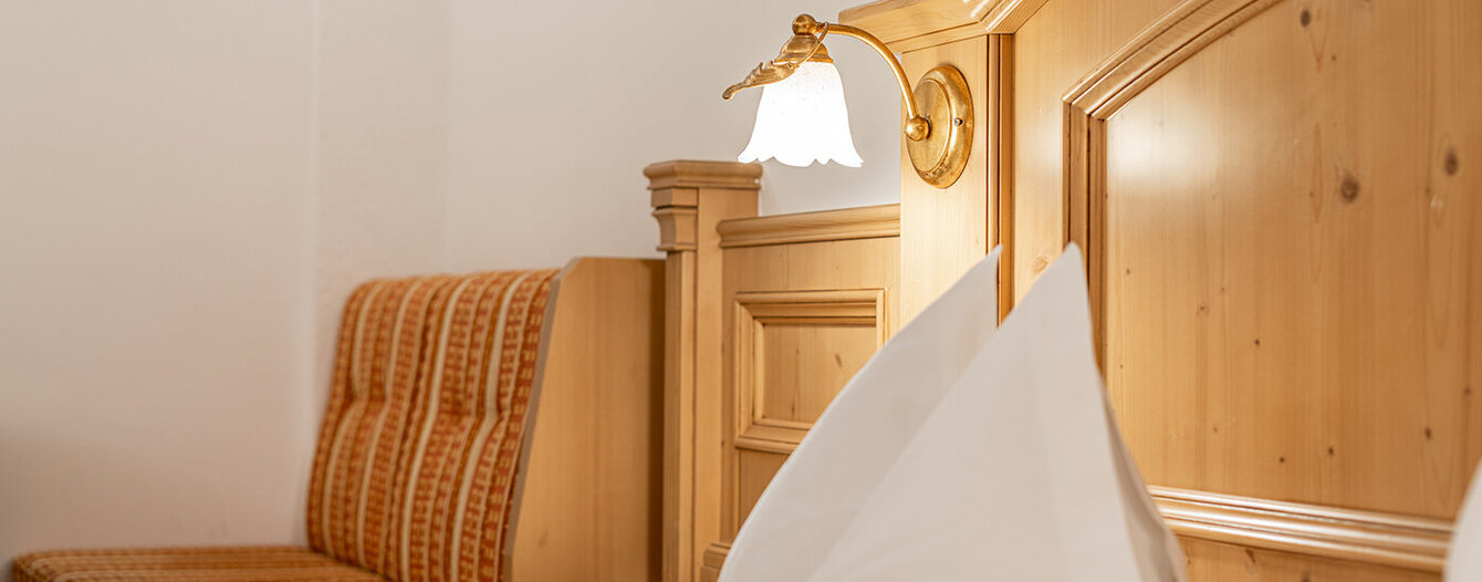 Nahaufnahme eines ordentlich gemachten Bettes mit weißen Kissen und Bettwäsche, daneben ein hölzernes Kopfteil und ein Nachttisch. Eine Wandlampe und eine gepolsterte Bank mit orangefarbenem Stoff sind ebenfalls zu sehen.