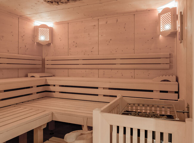 Ein geräumiger hölzerner Saunaraum mit gestuften Bänken an den Wänden, sanfter Beleuchtung in den oberen Ecken und einem beheizten Steinofen im Vordergrund. Der Innenraum hat eine warme, saubere und einladende Atmosphäre.