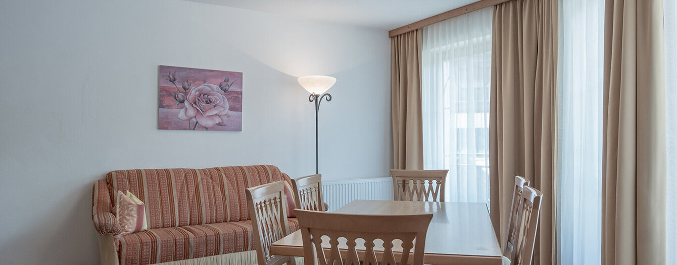 Ein helles Wohnzimmer mit einem gestreiften Sofa, einem rechteckigen Esstisch aus Holz mit vier Stühlen, einer Stehlampe, großen Fenstern mit beigen Vorhängen und einem Blumengemälde an der Wand.