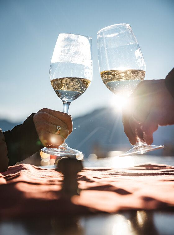 Zwei Personen stoßen im Freien im Sonnenlicht mit Weingläsern an, mit einem malerischen Berghintergrund und einem Sonnenflackereffekt.
