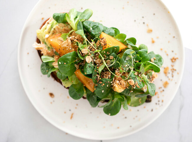 Offenes Sandwich mit Blattgemüse, gelben Tomatenscheiben und gestreuten Samen auf einem weißen Teller, von oben auf einer hellen Fläche fotografiert.