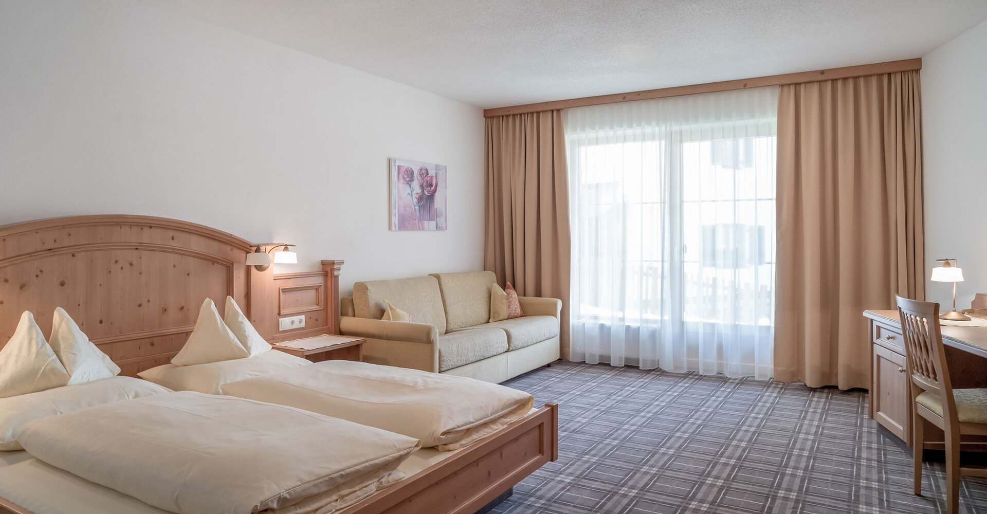 Ein Apartmentzimmer mit einem Doppelbett, einem beigen Sofa, einem Schreibtisch mit Stuhl und Lampe und großen Fenstern mit bodenlangen Vorhängen, die natürliches Licht hereinlassen. Die Einrichtung ist in hellem Holz und neutralen Tönen gehalten.