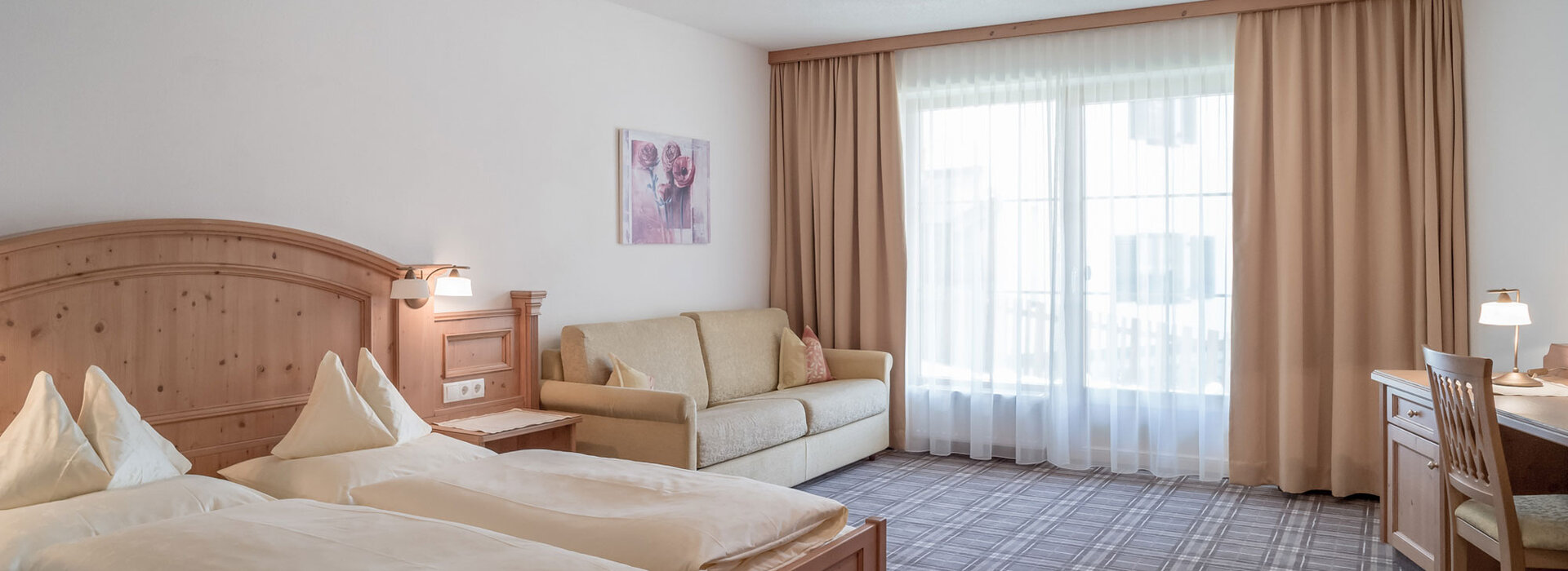 Ein Apartmentzimmer mit einem Doppelbett, einem beigen Sofa, einem Schreibtisch mit Stuhl und Lampe und großen Fenstern mit bodenlangen Vorhängen, die natürliches Licht hereinlassen. Die Einrichtung ist in hellem Holz und neutralen Tönen gehalten.