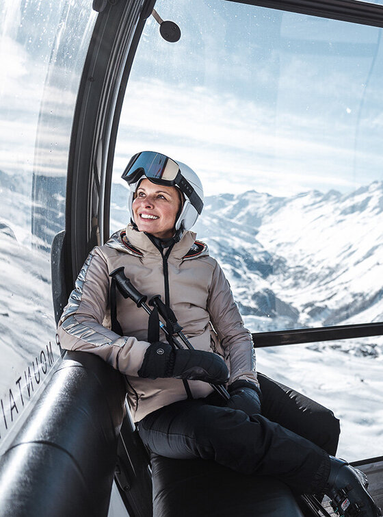 Zwei Skifahrer in Winterausrüstung sitzen in einer Gondel, hinter ihnen sind durch die Fenster verschneite Berge zu sehen.