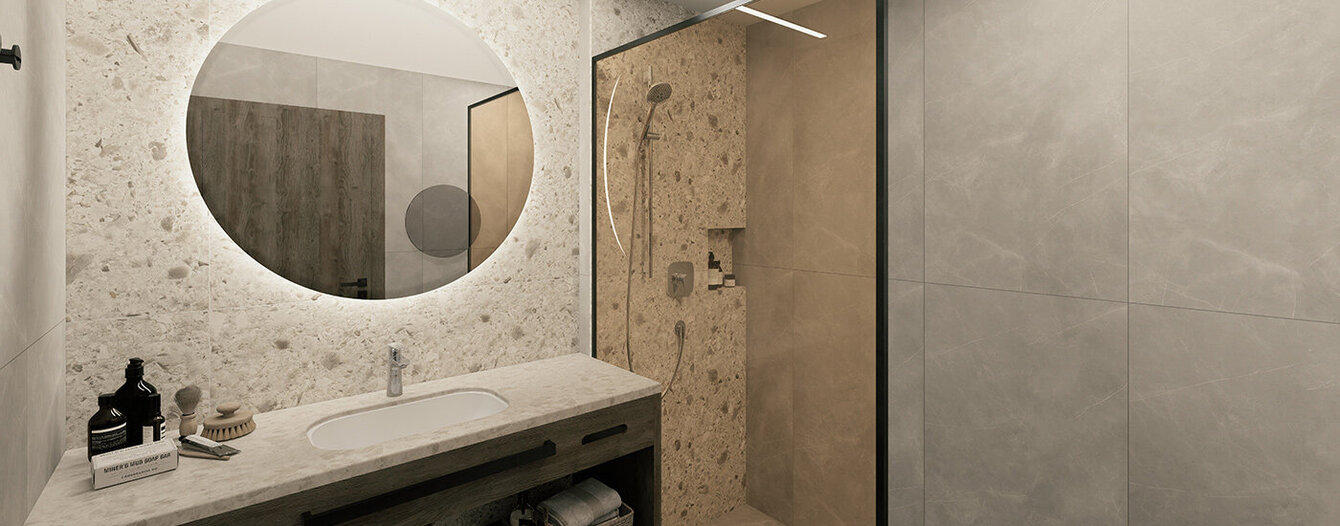 Modernes Badezimmer mit einem runden, hinterleuchteten Spiegel über einem Waschbecken und einem Waschtisch. Auf dem Regal liegen gerollte Handtücher, und auf der rechten Seite befindet sich eine Dusche mit Glasabdeckung und beigen Steinwänden. Das Gesamtdesign ist minimalistisch und neutral.