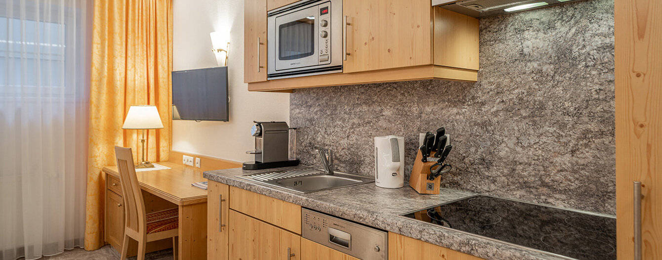 Eine kompakte Küche mit Holzschränken, einem Herd, einer Spüle, einer Mikrowelle, einem Geschirrspüler, einem Wasserkocher, einer Kaffeemaschine und einem Messerblock. Ein Schreibtisch mit Stuhl, Lampe und Wandfernseher steht neben einem Fenster mit gelben Vorhängen.