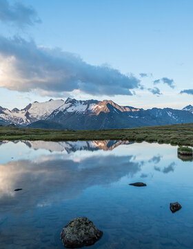 Schneebedeckte Berge und Wolken spiegeln sich in einem ruhigen See, umgeben von grasbewachsenem Gelände unter blauem Himmel.