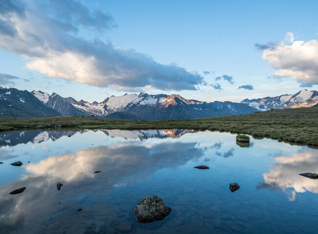 Schneebedeckte Berge und Wolken spiegeln sich in einem ruhigen See, umgeben von grasbewachsenem Gelände unter blauem Himmel.