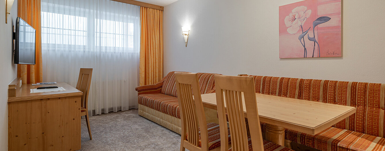 Ein gemütliches Wohnzimmer mit einem Holztisch, gestreiften Polstersesseln, einem orangefarbenen Sofa, einem an der Wand montierten Fernseher auf einem Holztisch und Vorhängen vor großen Fenstern. An der weißen Wand hängt ein geblümtes Gemälde.