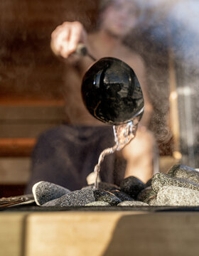 Eine Person gießt Wasser aus einer Schöpfkelle auf heiße Saunasteine, wodurch Dampf entsteht. Das Bild ist auf die Kelle und die Steine fokussiert, während die Person im Hintergrund verschwommen ist.