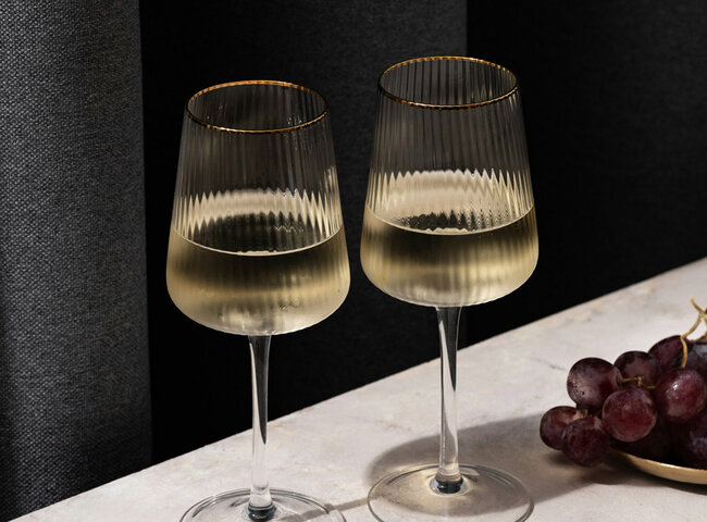 Zwei Gläser mit Weißwein auf einer hellen Fläche, daneben rote Weintrauben und Weichkäsestücke auf kleinen Tellern; im Hintergrund dunkle Vorhänge.