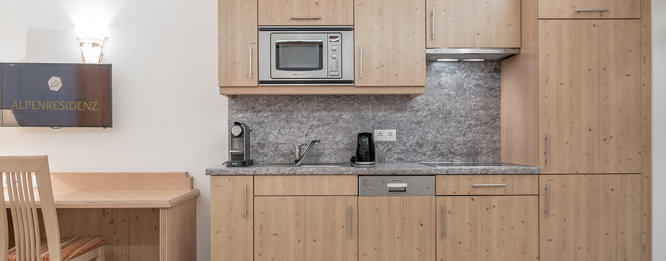 Eine kompakte Küchenzeile mit hellen Holzschränken, einer Mikrowelle, einem Herd, einem Geschirrspüler, einer Spüle und einer Kaffeemaschine. Auf der linken Seite stehen ein Schreibtisch und ein Stuhl, und im Vordergrund ist ein gefliester Boden zu sehen.