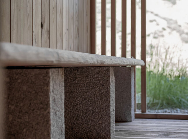 Eine Nahaufnahme einer minimalistischen Holzbank mit Steinauflagen, die auf einer Holzterrasse neben vertikalen Holzlatten steht und einen Blick auf Grünflächen und eine verschwommene Landschaft im Freien bietet.