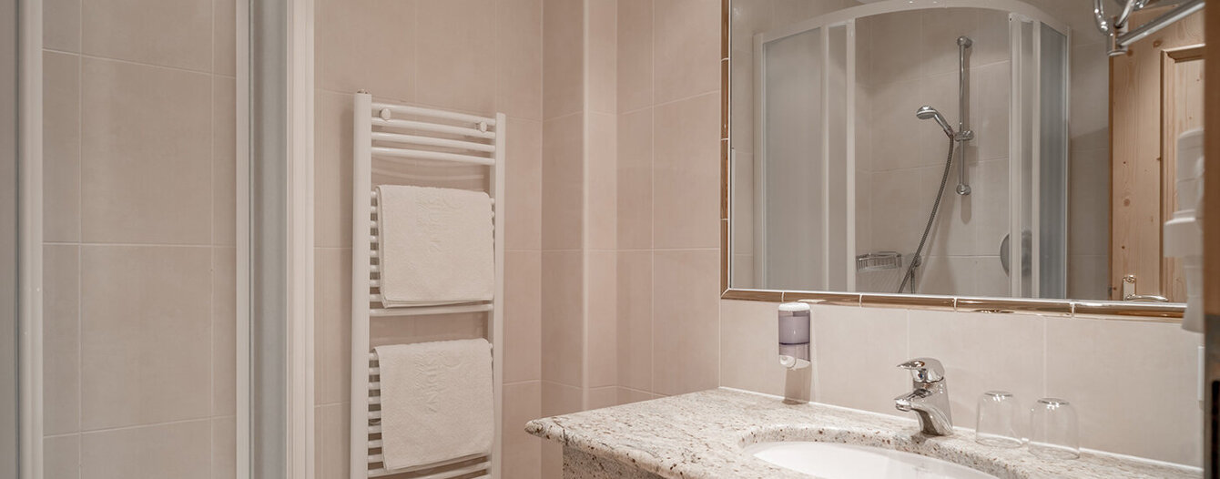 Modernes Badezimmer mit beigen Fliesen, einem großen Spiegel über einem Waschbecken mit Granitplatte, zwei Gläsern, einem Seifenspender, einem Handtuchhalter mit weißen Handtüchern und einer Glasduschkabine im Hintergrund.