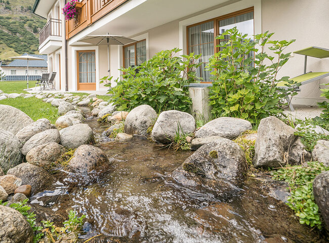 Ein kleiner Bach fließt über Felsen neben einem Gebäude, dessen Balkone mit bunten Blumen geschmückt sind; im Hintergrund sind Berge und ein teilweise bewölkter Himmel zu sehen.