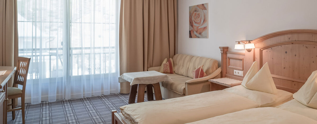 Ein gemütliches Zimmer mit einem Doppelbett, hellen Holzmöbeln, einem kleinen Sofa, einem Tisch, einem Stuhl und einem großen Fenster mit beigefarbenen Vorhängen und transparenten Vorhängen. Zur Ausstattung gehören ein Rosengemälde und ein karierter Teppichboden.