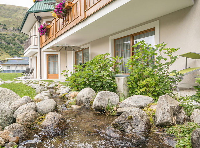 Ein kleiner Bach fließt über Felsen vor einem Gebäude mit Balkonen, die mit bunten Blumen geschmückt sind, in einer bergigen Landschaft unter einem teilweise bewölkten Himmel.