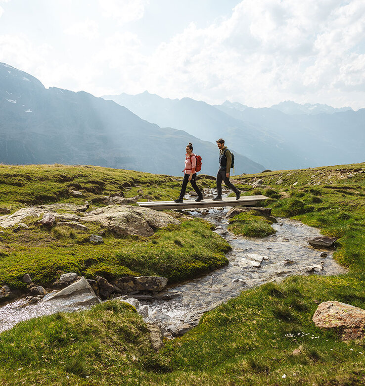 Zwei Personen wandern über eine kleine Holzbrücke über einen Bach in einer grasbewachsenen Berglandschaft mit fernen Gipfeln.