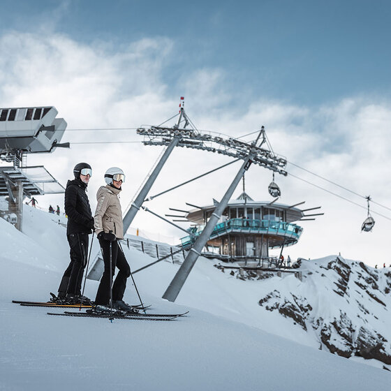 Zwei Personen in Skiausrüstung stehen auf einer verschneiten Piste in der Nähe einer Skiliftstation, mit Gondeln und einem modernen Gebäude im Hintergrund vor einem teilweise bewölkten Himmel.