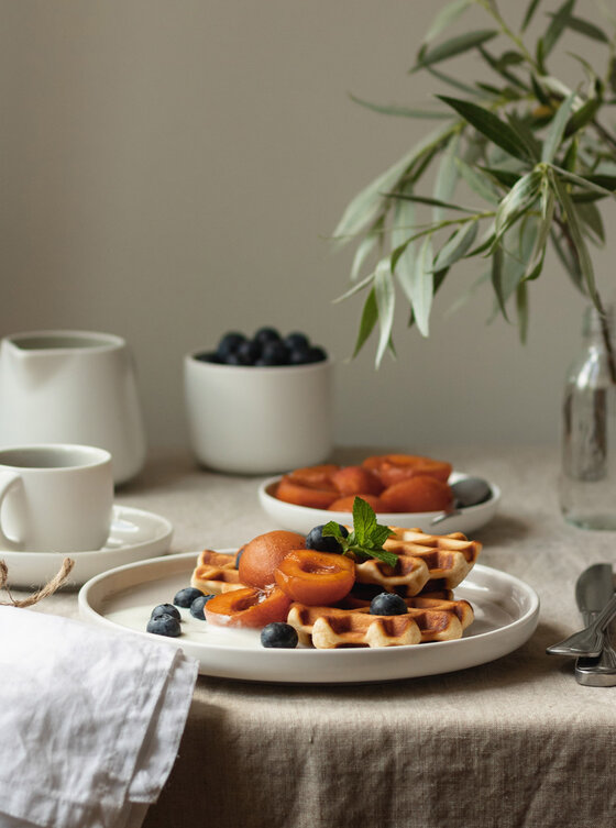 Ein gedeckter Frühstückstisch mit Waffeln, die mit Aprikosen, Blaubeeren und Minze auf einem weißen Teller belegt sind. Eine Tasse, ein kleiner Krug, eine Schale mit Blaubeeren und eine Glasvase mit Grünzeug stehen ebenfalls auf dem beigen Tischtuch.