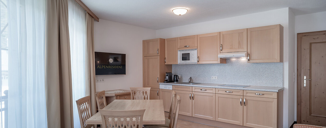 Eine gemütliche Küche und ein Essbereich mit hellen Holzschränken, einem Esstisch mit vier Stühlen, einem an der Wand montierten Fernseher, kariertem Fußboden und großen Fenstern mit beigen Vorhängen.