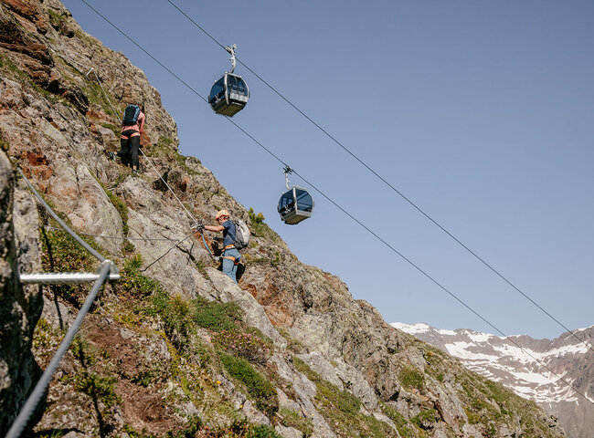 Zwei Kletterer mit Helmen und Rucksäcken steigen an einem gesicherten Seil einen felsigen, steilen Berghang hinauf. Drei Seilbahnen fahren gegen den klaren Himmel, im Hintergrund sind die schneebedeckten Gipfel zu sehen.