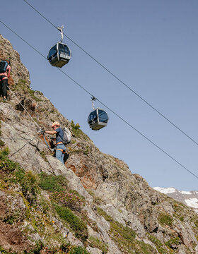 Zwei Kletterer mit Helmen und Rucksäcken steigen an einem gesicherten Seil einen felsigen, steilen Berghang hinauf. Drei Seilbahnen fahren gegen den klaren Himmel, im Hintergrund sind die schneebedeckten Gipfel zu sehen.