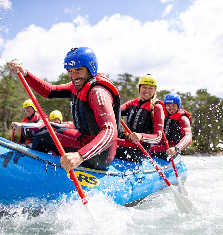 Eine Gruppe von Menschen, die Helme und Schwimmwesten tragen, paddelt lächelnd in einem blauen Schlauchboot, der Area 47, auf einem von Bäumen und Bergen umgebenen Fluss unter einem teilweise bewölkten Himmel.