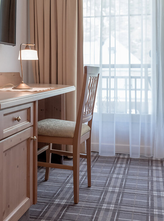 Ein hölzerner Schreibtisch mit einem Stuhl steht an einem Fenster mit durchsichtigen Vorhängen, durch die natürliches Licht einfällt. Eine Lampe und ein Flachbildfernseher befinden sich auf oder über dem Schreibtisch. Das Zimmer ist in neutralen Tönen gehalten und hat einen karierten Teppich.