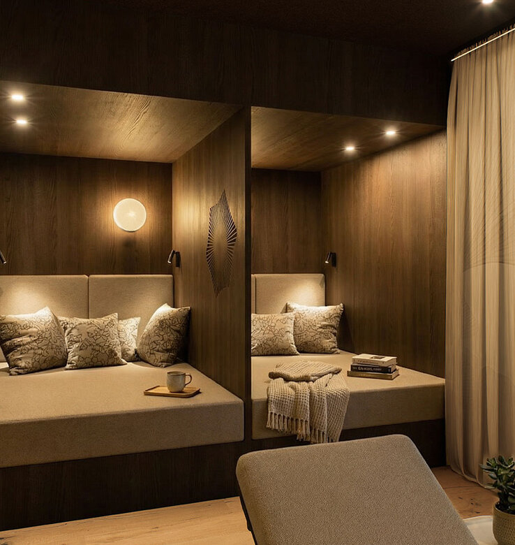 Ein gemütlicher, moderner Entspannungsraum mit zwei eingebauten Sitzecken, die jeweils mit Kissen, Überwürfen und Decken ausgestattet sind. Die warme Beleuchtung und die Holzwände schaffen eine ruhige Atmosphäre.
